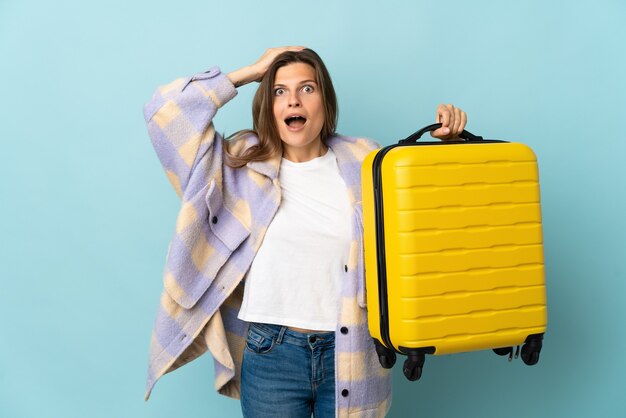 Young Slovak woman isolated on blue background in vacation with travel suitcase and surprised