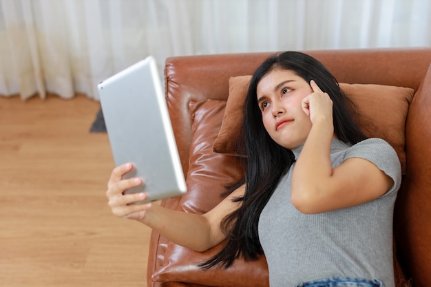 Young smart and active asian female lying on sofa in living room, using tablet while thinking something, Lifestyle and technology