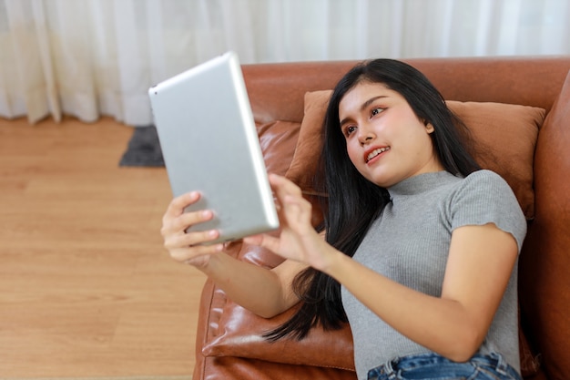 Young smart and active asian female lying on sofa in living room, using tablet with happy smiling face, Lifestyle and technology