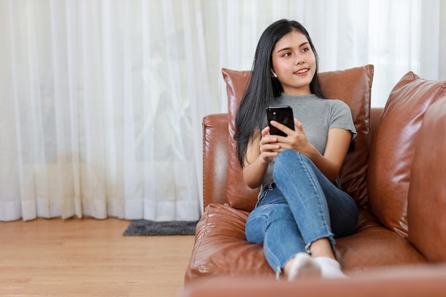 Young smart and active asian female sitting on sofa in living room, using smartphone with happy smiling face