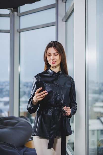 Young smart businesswoman in casual clothes working with phone while standing near window and reporting and writing notes and against blurred modern office interior