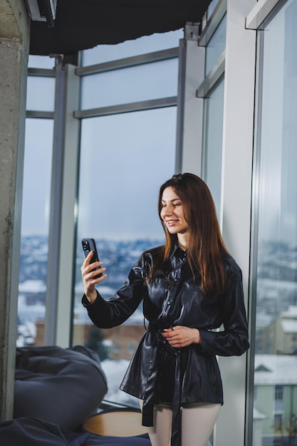 Young smart businesswoman in casual clothes working with phone while standing near window and reporting and writing notes and against blurred modern office interior