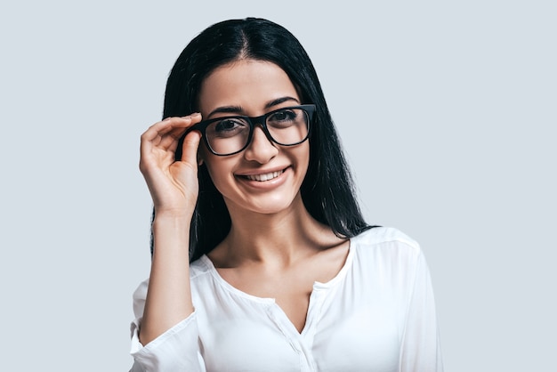 Young and smart. Young attractive woman in white shirt and glasses smiling while standing against grey background