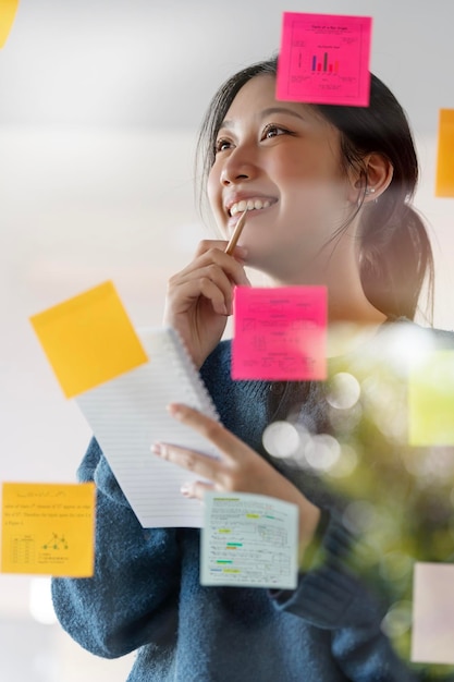 Young smiley attractive businesswoman using post it notes in glass wall to writing strategy business plan to development grow to success