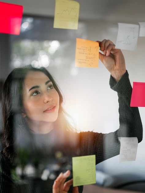 Young smiley attractive businesswoman using post it notes in glass wall to writing strategy business plan to development grow to success