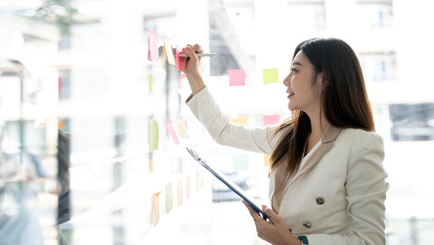 Young smiley attractive businesswoman using post it notes in glass wall to writing strategy business plan to development grow to success