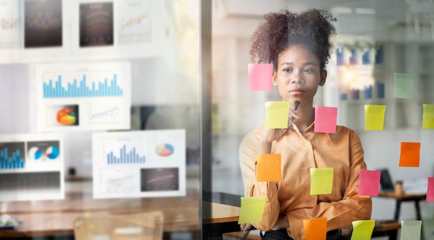 Young smiley attractive businesswoman using sticky notes in glass wall to writing strategy business plan to development grow to success