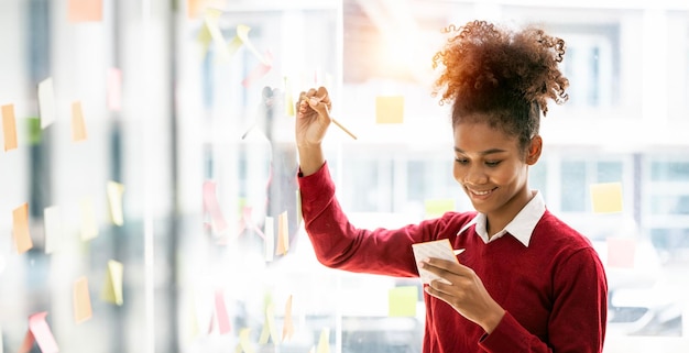 Young smiley attractive businesswoman using sticky notes in glass wall to writing strategy business plan to development grow to success