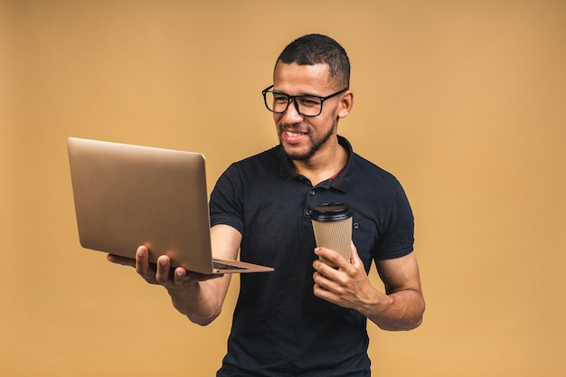 Young smiling african american black man standing and using laptop computer isolated over beige background Drinking coffee