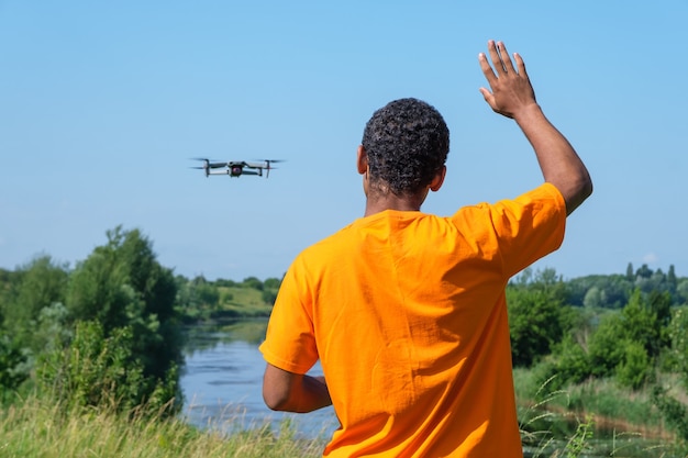 Young smiling African American man operating drone with controller standing with back and waving with his hand on the meadow near the river.