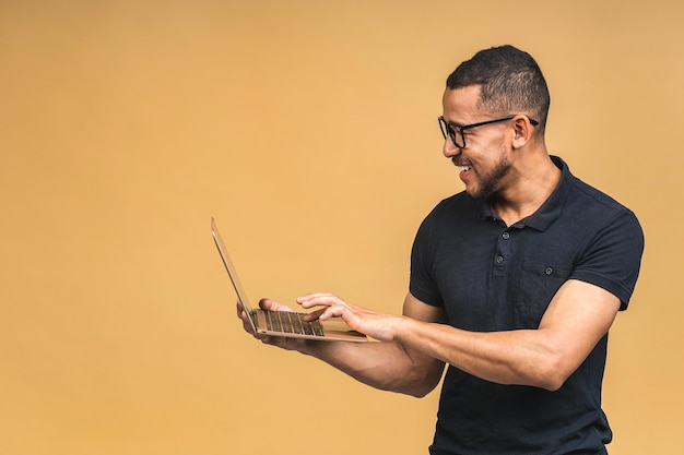 Young smiling african american man standing and using laptop computer isolated over beige background