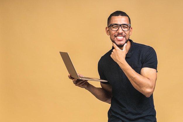 Young smiling african american man standing and using laptop computer isolated over beige background