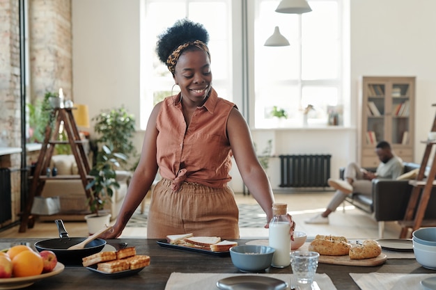 Young smiling African woman in casualwear standing by electric stove and taking bottle of milk while cooking dinner for her family in the kitchen