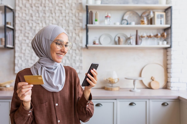 Young smiling arab woman in hijab using phone and credit card at home in kitchen do online shopping