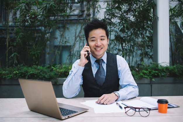 Young smiling Asian businessman talking on phone while sitting at desk with laptop