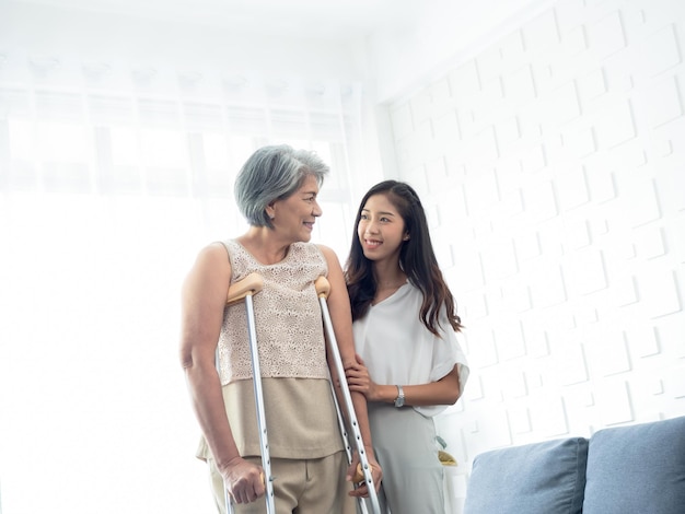 Young smiling Asian females carefully hold and support the arms of elderly women trying to walk on crutches in recovery room helping old women health care therapy senior patient at home concept