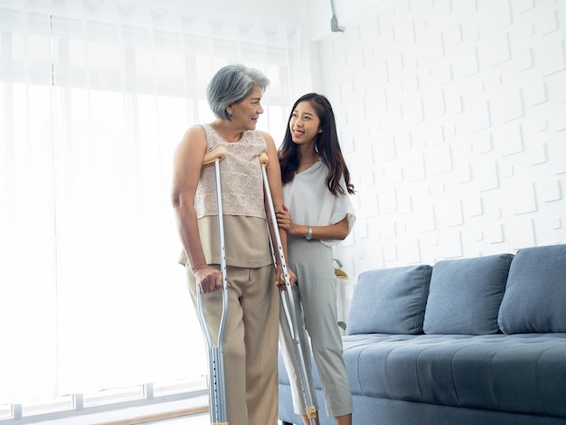 Young smiling Asian females carefully hold and support the arms of elderly women trying to walk on crutches in recovery room helping old women health care therapy senior patient at home concept