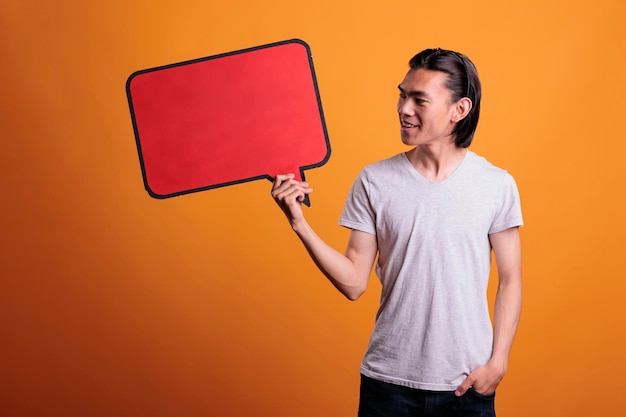 Young smiling asian man holding empty red speech bubble with copy space, advertising product. Cheerful teenager standing, looking at blank dialog frame with place for text