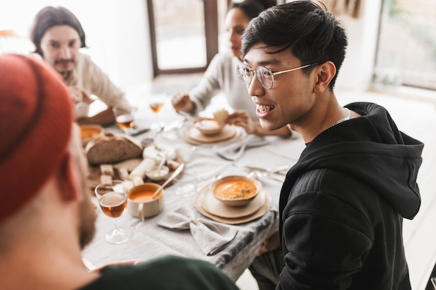 Young smiling asian man with dark hair in eyeglasses and hoodie sitting at the table happily 