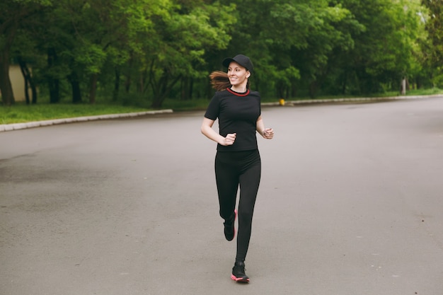 Young smiling athletic beautiful brunette woman in black uniform, cap training doing sport exercises, running, jogging, looking aside on path in city park outdoors