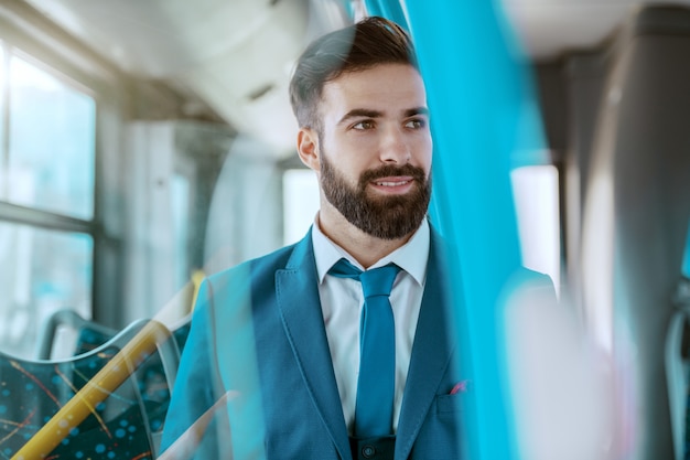 Young smiling attractive businessman in blue suit sitting in public bus and looking away.