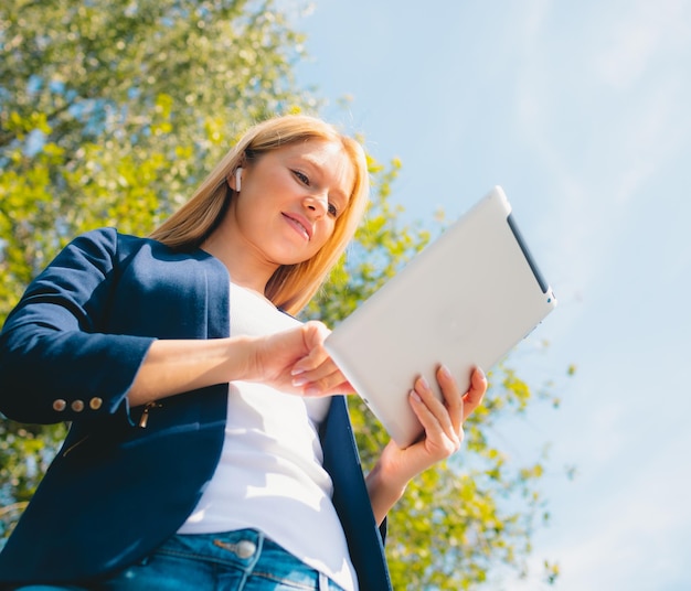 Young smiling beautiful woman in park with tablet gadget and wireless headphones enjoying green energy concept remote work