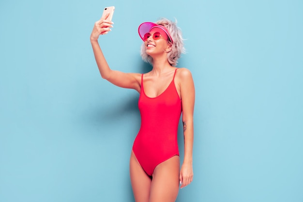 Young smiling blond model in summer swimwear red bathing suit and transparent visor cap Sexy carefree woman having fun and going crazy Female posing near blue wall in studio Taking selfie