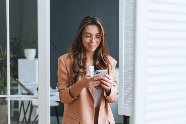 Young smiling brunette business woman in glasses with long hair in stylish beige suit using smartphone in the modern office