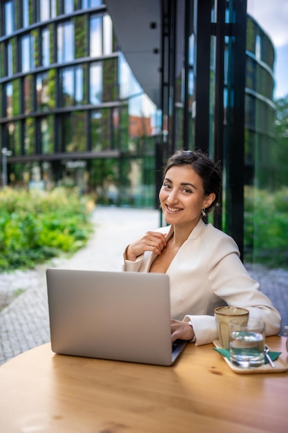 Young smiling brunette business woman in stylish smart dress working on laptop in cafe at street of