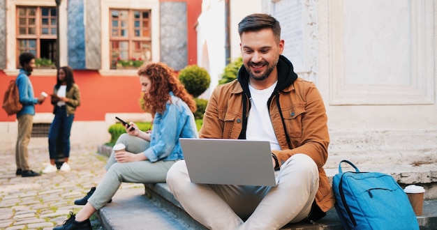 Young smiling brunette man wearing casual clothes using laptop computer outdoors in the street while...