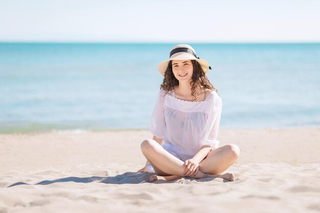 Young smiling brunette teenager girl sittin on the beach near the sea Happy Caucasian woman on vacation at sunny day on seashore