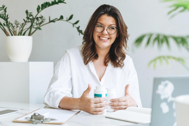 Young smiling brunette woman nutritionist plus size in white shirt working at laptop on table with house plant in the bright modern office Doctor communicates with the patient online