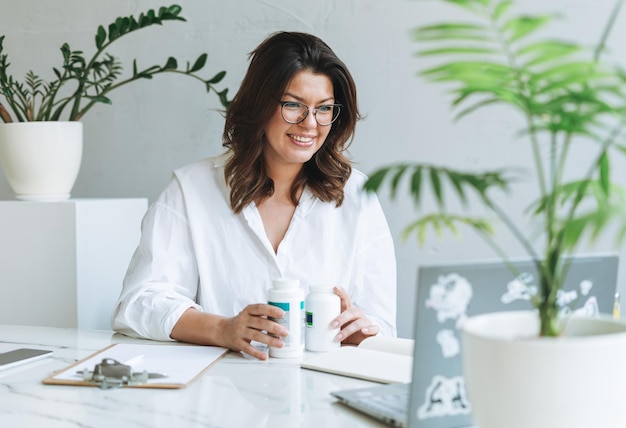 Young smiling brunette woman nutritionist plus size in white shirt working at laptop on table with house plant in the bright modern office Doctor communicates with patient online