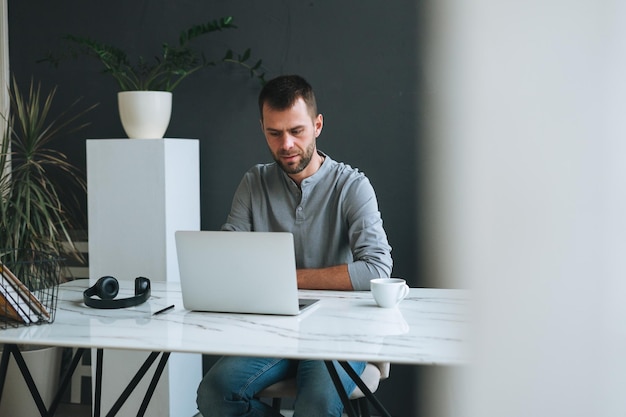 Young smiling business man in casual grey longsleeve working at laptop in dark modern office room