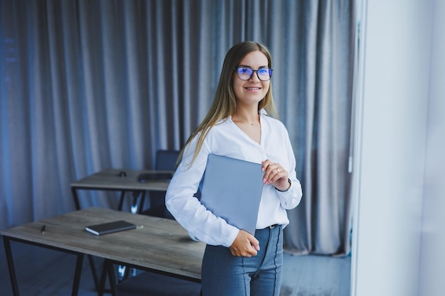 A young smiling business woman is standing in the office by the window with a laptop in her hands Woman manager in glasses and white shirt