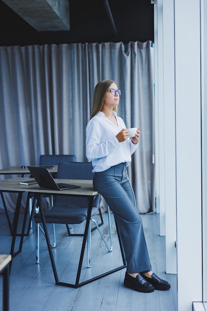 Young smiling business woman standing in office near window with coffee in hands A female manager in glasses and a white shirt is resting from work