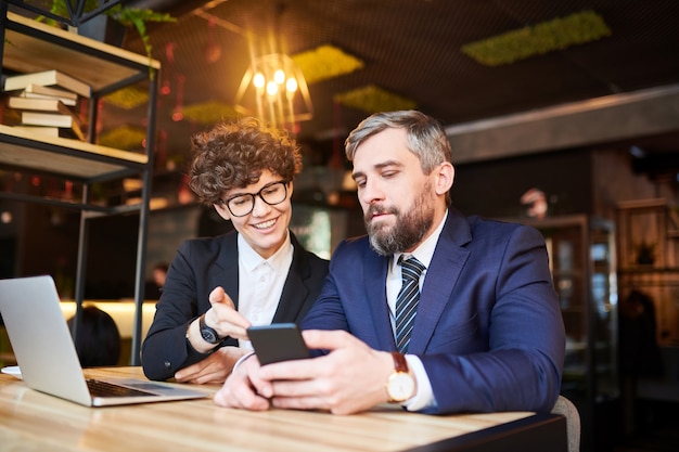 Young smiling businesswoman showing her colleague data in his smartphone during discussion of working points