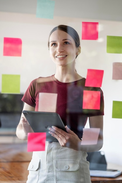 Young smiling caucasian businesswoman mentor leader write tasks creative ideas on sticky post it notes on glass wall