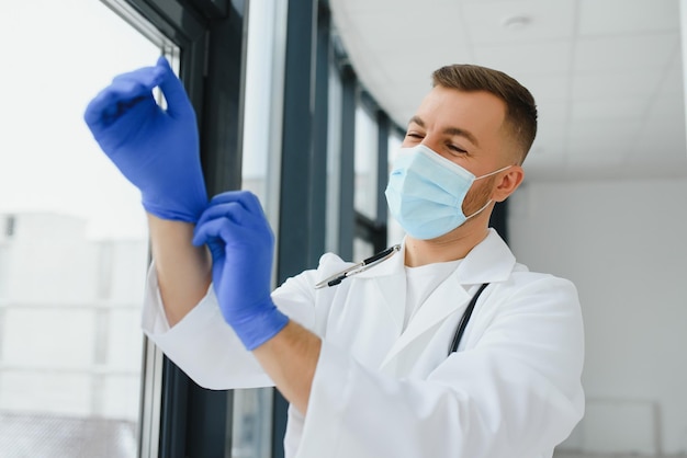 Young smiling Caucasian male doctor in white uniform with eyeglasses and stethoscope around neck putting rubber gloves before patient examination