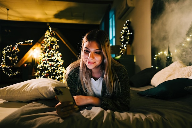 Young smiling caucasian woman using mobile phone in holidays at home on the bed.