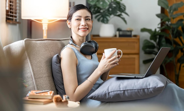 Young smiling confident asian woman relaxing at home listening music via laptop computer and enjoying with hot drink