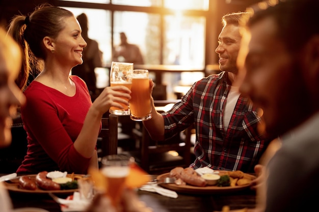 Young smiling couple celebrating and toasting with beer while having lunch with friends in a pub