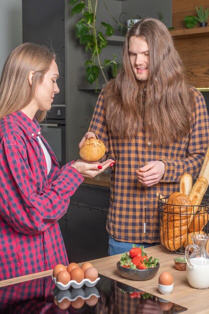 Young smiling couple in checkered shirts in kitchen longhaired man giving bread to his girlfriend