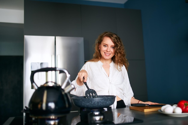 Young smiling curly caucasian woman cooks breakfast on a frying pan young adult female making