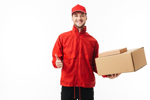 Young smiling delivery man in red cap and jacket holding boxes in hand happily showing thumb up gesture while 