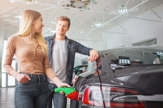 Young smiling family couple buying first electric car in the showroom. Close-up of attractive woman charging ecological hybrid car with the power cable supply plugged in while watching at her husband.