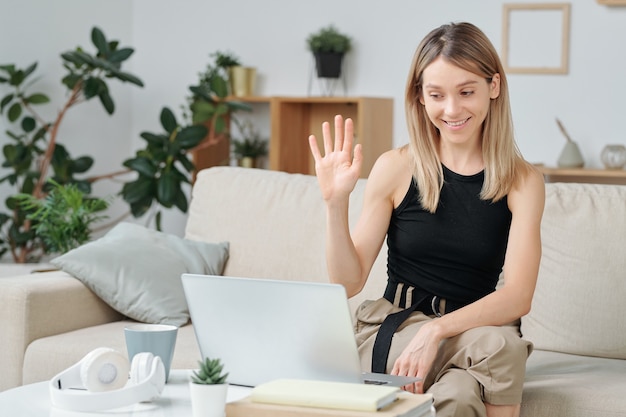 Young smiling female in casualwear sitting on sofa and waving hand to her friend in video chat while looking at laptop display and saying hello