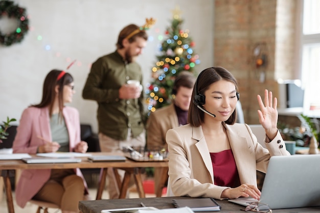 Young smiling female customer support representative with headset looking at laptop display and waving hand while greeting client