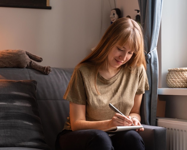 Young smiling female taking notes in journal. Writing down ideas and thoughts in secret diary. Planning and making lists. Expecting and waiting in cozy atmosphere