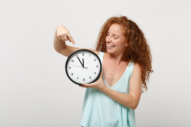 Young smiling funny redhead woman girl in casual light clothes posing isolated on white wall background, studio portrait. People lifestyle concept. Mock up copy space. Pointing index finger on clock.